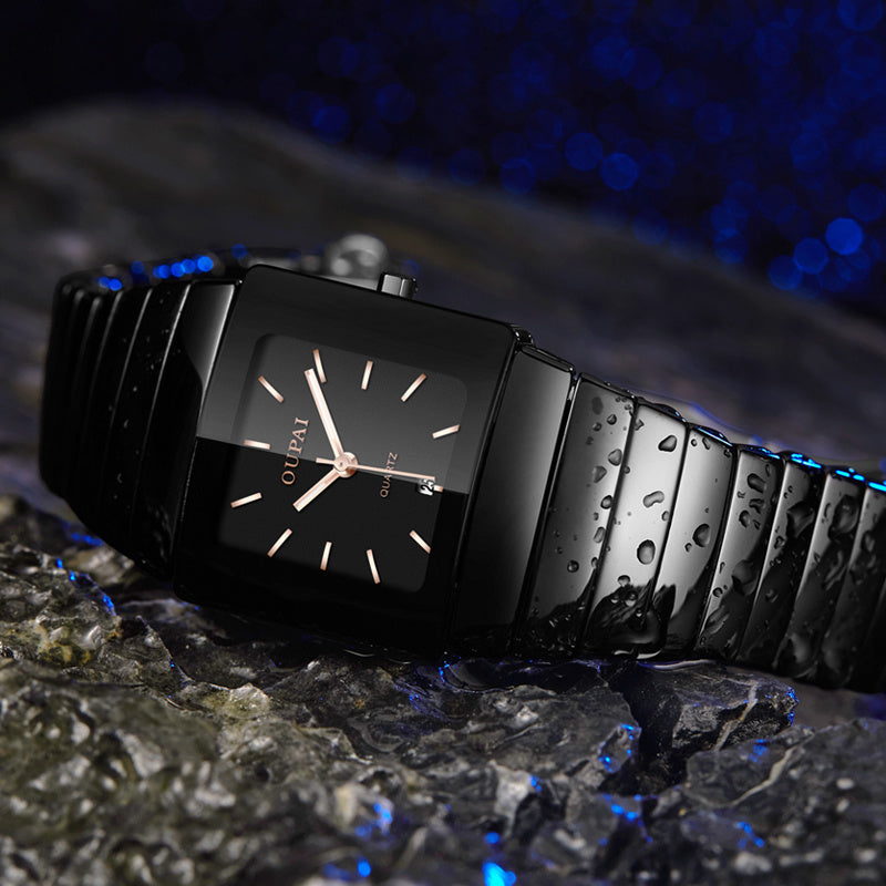 Black square-faced wristwatch with a black linked bracelet and light-colored hands, photographed with water droplets on a textured surface.