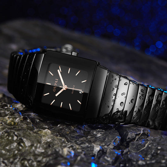 Black square-faced wristwatch with a black linked bracelet and light-colored hands, photographed with water droplets on a textured surface.